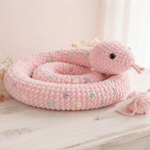 May include: A coiled plush snake toy made of soft pink yarn with a repeating pattern of small pastel hearts. The snake features black plastic eyes and a bifurcated tongue. A small ball of matching pink yarn and a white crochet hook are nearby.