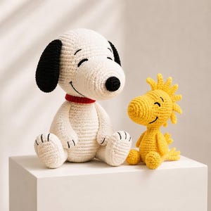 Puede incluir: Un muñeco beagle de ganchillo blanco con orejas negras, un collar rojo y una nariz negra sentado junto a un muñeco pájaro de ganchillo amarillo más pequeño. Ambas figuras tienen ojos cerrados bordados y bocas sonrientes.