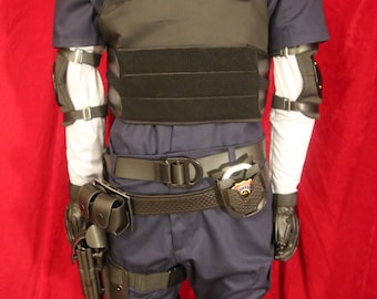 Può includere: Un set di uniforme tattica composto da un gilet nero con scritta bianca R.P.D., una camicia blu scuro a maniche corte, maniche lunghe bianche, cintura tattica nera con fondina e manette, guanti neri e ginocchiere nere su pantaloni blu scuro.