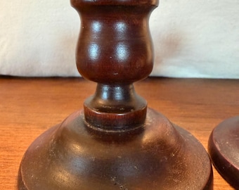 May include: A dark brown wooden candle holder with a smooth, polished finish. The object features a wide, circular base that tapers upwards into a rounded, bulbous stem, topped with a flat, circular platform designed to hold a candle.