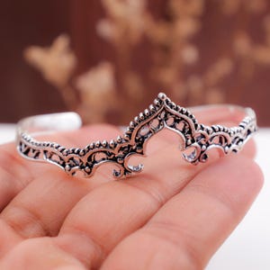 May include: A silver-toned metal cuff bracelet featuring an intricate, openwork filigree design with a pointed, arched centre peak. The metal has a darkened, oxidised finish in the recessed areas to highlight the detailed scrollwork patterns.