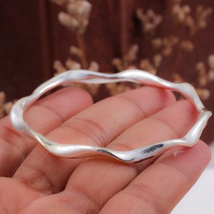 May include: A silver-toned bangle bracelet featuring a continuous, undulating wavy design. The metal surface has a smooth, polished finish that reflects light along its curved, organic shape, creating a fluid and rhythmic appearance throughout the band.