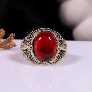 May include: A ring featuring a large, oval-shaped, deep red cabochon stone set in a metallic band with an antique gold finish. The band displays intricate, textured relief patterns resembling stylised feathers or wings on both sides of the stone.