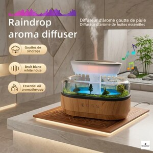 Peut inclure: Un diffuseur d'arôme goutte de pluie avec une base en imitation bois et une partie supérieure transparente contenant un paysage miniature avec des arbres et de l'eau. L'appareil émet une brume et affiche le texte Raindrop aroma diffuser, Gouttes de sindrops, Bruit blanc white noise et Essential oil aromatherapy.
