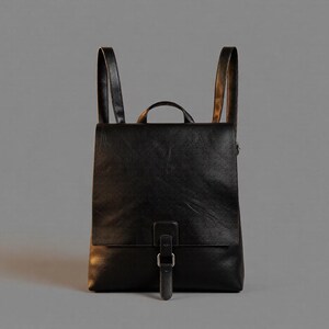 May include: A black backpack with a smooth texture, featuring a large front flap that covers the main compartment. The bag includes a top handle, two vertical shoulder straps, and a central vertical strap secured with a silver-toned metal buckle.