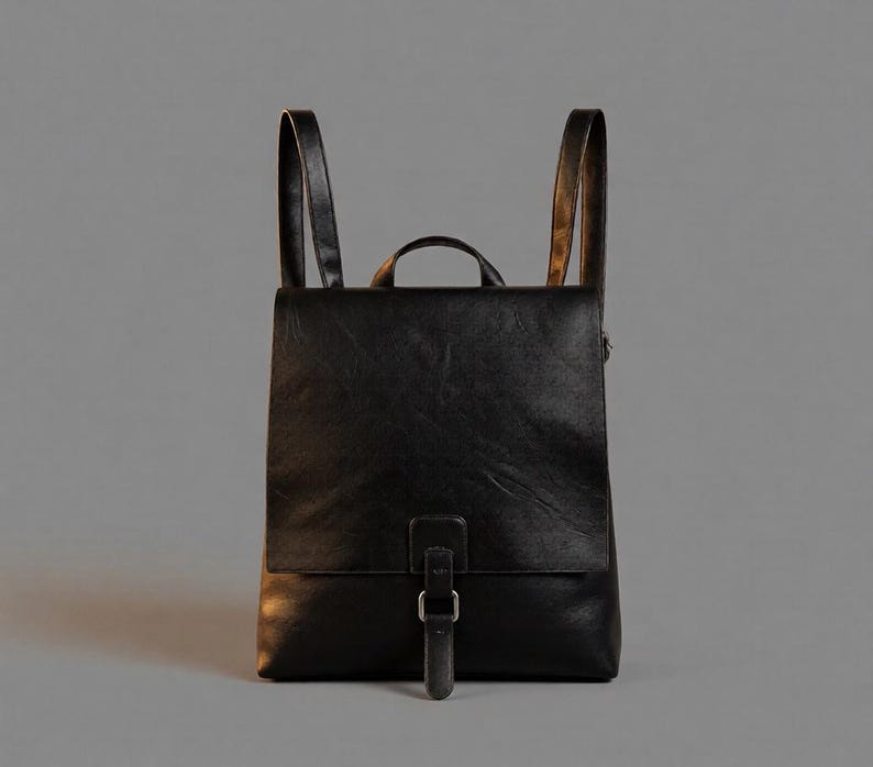May include: A black backpack with a smooth texture, featuring a large front flap that covers the main compartment. The bag includes a top handle, two vertical shoulder straps, and a central vertical strap secured with a silver-toned metal buckle.