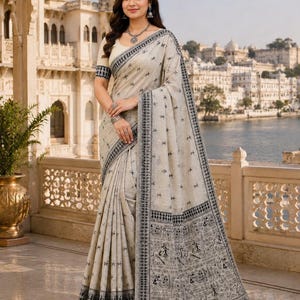 May include: A beige saree featuring a black printed pattern of small motifs across the fabric and a wide border with detailed black illustrations of figures and geometric shapes. The garment includes a matching short-sleeved blouse with black trim.