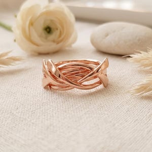 Op de afbeelding: Een roségouden ring met een complex, verweven ontwerp van meerdere dunne, gepolijste metalen banden die elkaar kruisen om een doorlopend, getextureerd en gelaagd uiterlijk te creëren met een gladde, reflecterende afwerking over het hele sieraad.