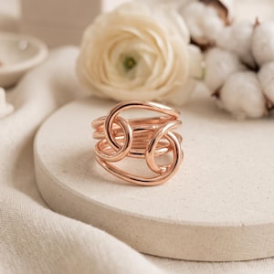 Puede incluir: Un anillo de oro rosa con un diseño de bucles interconectados de varias capas. La banda consta de varios hilos de metal pulido y curvado que se superponen para formar una forma fluida y continua, creando una apariencia compleja y estratificada.
