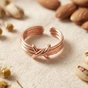 Op de afbeelding: Een roségoudkleurige ring met een ontwerp van vier parallelle draden die in het midden kruisen en in elkaar verstrengeld zijn. De ring heeft een open constructie en een gladde, gepolijste metalen afwerking over de gehele band.