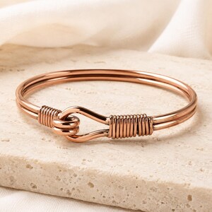 Puede incluir: Un brazalete de tono oro rosa con un diseño de doble alambre. La pulsera incluye un mecanismo de cierre tipo gancho donde los extremos de los alambres están asegurados con un enrollado de alambre apretado, creando un acabado metálico pulido.