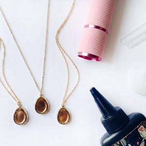 May include: Three gold-toned necklaces with oval-shaped pendants, a pink cylindrical tool with a textured grip, two clear plastic stirrers, a white circular object, and a black squeeze bottle with a label featuring an owl and leaf pattern.