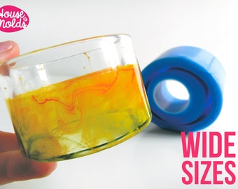 May include: A clear glass container filled with yellow liquid with orange swirls. The container is being held by a hand. A blue silicone mould is in the background. The text "WIDE SIZES" is in pink letters on a white background.