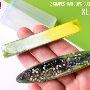 Peut inclure: Moule en silicone transparent pour la fabrication de deux formes de pinces à cheveux. Le moule est présenté avec deux pinces à cheveux finies, une rectangulaire et une ovale. La pince à cheveux ovale est décorée d'étoiles scintillantes colorées. Le moule est étiqueté "2 SHAPES HAIR CLIPS CLEAR MOLD XL SIZE".