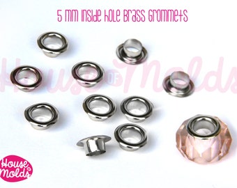 May include: A close-up of 5 mm inside hole brass grommets. The grommets are silver in color and are arranged in a scattered pattern on a white surface. There is a pink crystal bead with a silver rim in the lower right corner of the image. The text "House of Molds" is visible in the image.