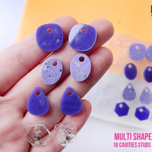 Könnte beinhalten: Lila und klare Harzohrringe in verschiedenen Formen, darunter Tropfen, Oval und Quadrat, mit Glitzer in einigen eingebettet. Die Ohrringe werden auf einer Hand und einem weißen Tablett mit dem Text "MULTI SHAPES MOLD 16 CAVITIES STUDS EARRINGS" gezeigt.