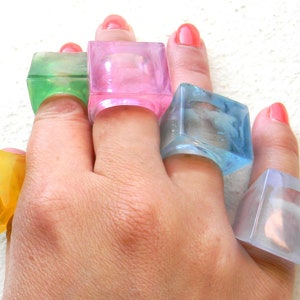May include: Five colorful square-shaped rings made of clear plastic. The rings are worn on the fingers of a hand. The rings are in shades of pink, green, blue, yellow, and clear.