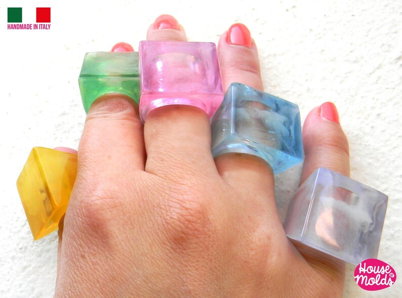 May include: Five colorful square-shaped rings made of clear plastic. The rings are worn on the fingers of a hand. The rings are in shades of pink, green, blue, yellow, and clear.
