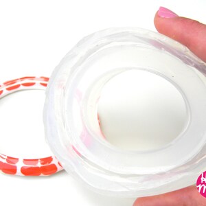 May include: A clear silicone mould for making a ring. The mould is made of two parts, and the top part is being held by a hand. The mould is designed to create a ring with a wide band and a raised centre.