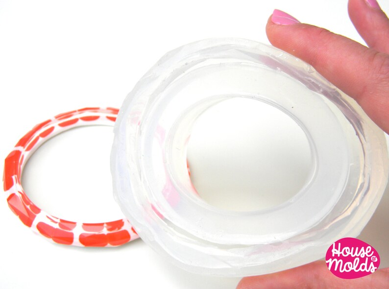 May include: A clear silicone mould for making a ring. The mould is made of two parts, and the top part is being held by a hand. The mould is designed to create a ring with a wide band and a raised centre.