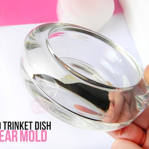 May include: Clear mould for a trinket dish. The mould is made of clear plastic and is shaped like a shallow bowl. The mould is being held in a hand. The text "BOLD TRINKET DISH CLEAR MOLD" is printed on a white background below the mould.