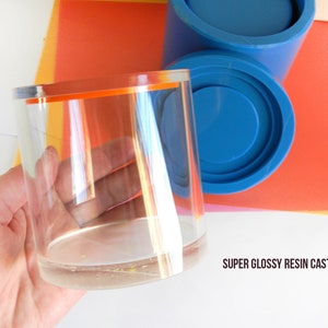 May include: A clear glass cylinder with an orange rim, held in a hand. The cylinder is on top of a blue silicone mould, with the text "SUPER GLOSSY RESIN CASTING" below.