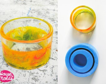 May include: Two silicone moulds for resin casting. One mould is blue and has a round shape with a smaller round opening inside. The other mould is yellow and orange and is a ring shape with a round opening inside. The moulds are used to create resin coasters or other decorative items.