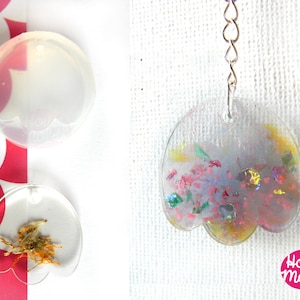 Puede incluir: Dos moldes de silicona transparentes en forma de flor. El molde de la izquierda tiene una flor seca en su interior. El molde de la derecha tiene una pieza de resina colorida en su interior con purpurina y flores secas. La pieza de resina está unida a una cadena plateada.