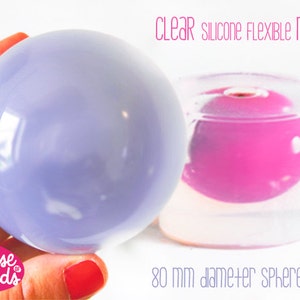 Peut inclure: Un moule sphérique en silicone transparent avec le texte "CLEAR SILICONE FLEXIBLE MOLD!" et "80 mm diameter SPHERE MOLD". Une sphère violet clair est tenue à côté du moule, qui contient une sphère rose.