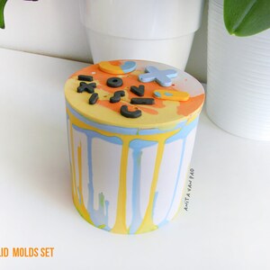 May include: A colourful, abstractly painted jar with lid mould set. The jar is decorated with yellow, blue and orange paint and has a variety of shapes and letters on the lid. The jar is labelled "Anita Van Pad" and "Handmade in Italy".