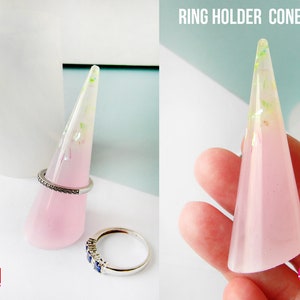 Può includere: Uno stampo in silicone trasparente per realizzare un cono porta anelli. Lo stampo ha la forma di un cono e presenta un effetto sfumato rosa e bianco. Lo stampo è mostrato con un cono porta anelli finito realizzato con resina e glitter.