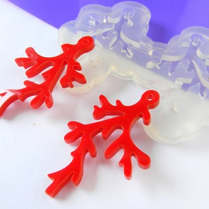 May include: Two red resin coral branch charms with a hole for hanging. The charms are next to a clear silicone mold of the same design. The mold is labeled "Handmade in Italy".