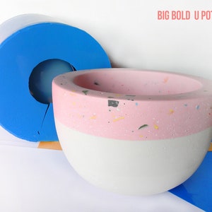 May include: A blue silicone mould for making a large, round concrete planter with a pink and white speckled finish. The mould is in two pieces and has a hole in the centre for drainage. The planter is labelled "BIG BOLD U POT MOLD" and "House Molds".