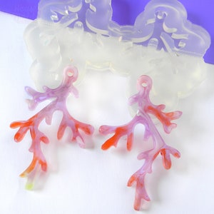 May include: Two coral-colored resin pieces shaped like branches with a hole at the top for hanging. The pieces are made in a clear mold, which is also visible in the image.