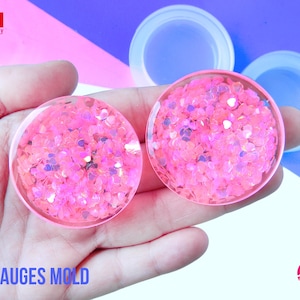 May include: Two pink silicone ear gauge molds with a heart-shaped glitter design. The molds are held in a hand against a pink and blue background. The text "EAR GAUGES MOLD" is at the bottom of the image. The text "House of Molds" is in a red circle at the bottom right of the image.