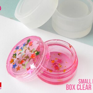 Peut inclure: Un moule rond en plastique transparent pour boîte avec un couvercle rose. Le moule est présenté avec une pièce en résine finie à l'intérieur. La pièce en résine est remplie de paillettes et d'étoiles. Le moule est étiqueté "SMALL ROUND BOX CLEAR MOLD".
