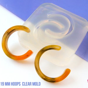 Könnte beinhalten: Transparente Silikonform zum Herstellen von 19 mm Creolen. Die Form ist mit zwei fertigen Ohrringen gezeigt, einem orangen und einem gelben.