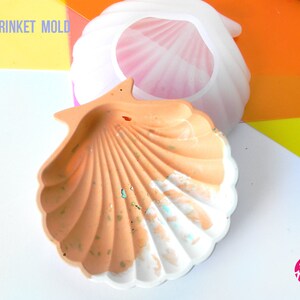 May include: A white silicone shell trinket mold with a pink interior. The mold is next to a finished, painted shell trinket with a peach and white color scheme. The text "SHELL TRINKET MOLD" is visible in the top left corner of the image. The text "House Molds" is visible in the bottom right corner of the image.
