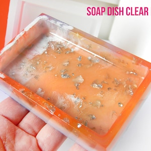 Op de afbeelding: Transparante zeepvorm met een kleurrijk, gelaagd ontwerp. De vorm is gevuld met een heldere hars met zilveren vlokken. De vorm wordt in een hand gehouden.