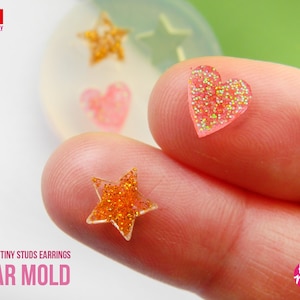 May include: Close-up of four clear silicone molds for making tiny stud earrings. The molds are shaped like hearts and stars. The molds are filled with glitter resin in pink and orange.