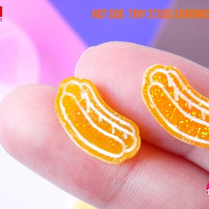 Puede incluir: Un par de pendientes en forma de hot dog de color naranja y blanco. Los pendientes están hechos de resina y tienen un acabado brillante. Los pendientes son pequeños y son perfectos para el uso diario.