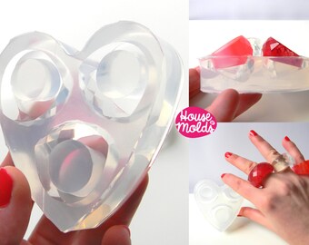 May include: A clear silicone mold shaped like a heart with three ring-shaped cavities. The mold is being held in a hand. The image also shows a finished ring made with the mold, which is red and clear.