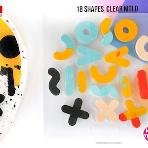 May include: A clear plastic mould with 18 different shapes. The shapes are filled with colourful resin and include circles, ovals, and abstract shapes. The mould is labelled "18 Shapes Clear Mould" and "Casting Made with ResinObite".