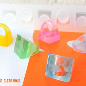May include: Four cube-shaped rings made with clear resin. The rings are in different colors: yellow, green, pink, and clear. The rings are sitting on a white surface with an orange surface in the background. The image also shows a clear silicone mold with four cavities for making the rings. The text "CUBE RINGS CLEAR MOLD" is at the bottom of the image. The text "House Molds" is at the bottom right of the image.
