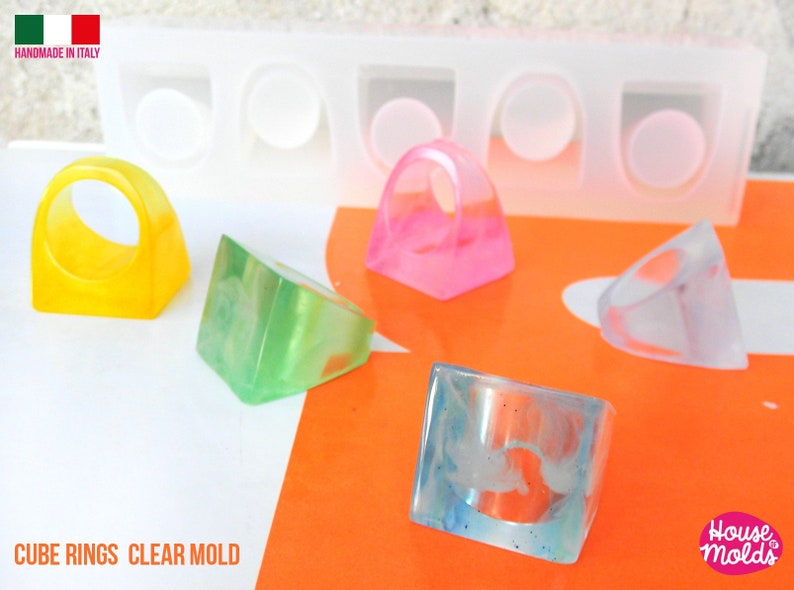 May include: Four cube-shaped rings made with clear resin. The rings are in different colors: yellow, green, pink, and clear. The rings are sitting on a white surface with an orange surface in the background. The image also shows a clear silicone mold with four cavities for making the rings. The text "CUBE RINGS CLEAR MOLD" is at the bottom of the image. The text "House Molds" is at the bottom right of the image.