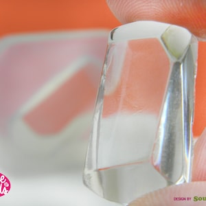 Peut inclure: Un moule transparent de forme géométrique pour la fabrication d'objets en résine. Le moule est tenu dans une main, et l'arrière-plan est une surface orange floue.