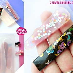 Puede incluir: Molde de silicona transparente para hacer dos formas de pinzas para el pelo. Un molde es un rectángulo con un diseño de media luna y el otro es una forma curva con un diseño de estrella. El molde se muestra con pinzas para el pelo terminadas.