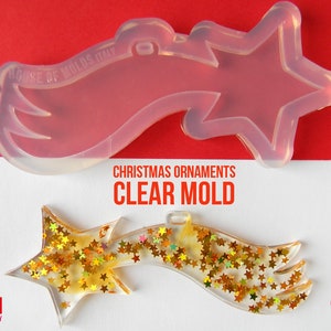 Puede incluir: Molde de silicona transparente para hacer adornos navideños en forma de estrella fugaz. El molde se muestra con un adorno terminado relleno de purpurina dorada.