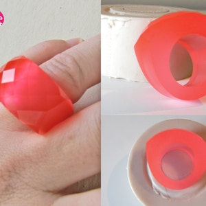 以下が含まれることがあります： ピンクの樹脂製のリングが2つ。1つはファセットデザイン、もう1つはシンプルな楕円形。リングは白い表面と指の上に置かれています。