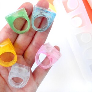 May include: Five colorful resin rings in a variety of shapes and sizes. The rings are made in a clear resin with different colors mixed in. The rings are displayed on a hand. The rings are made using a silicone mold that is also shown in the image. The mold has three different ring shapes. Handmade in Italy.